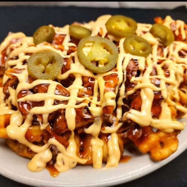 Waffle fries loaded with BBQ chicken, creamy sauce, and sliced jalapeños on a white plate.